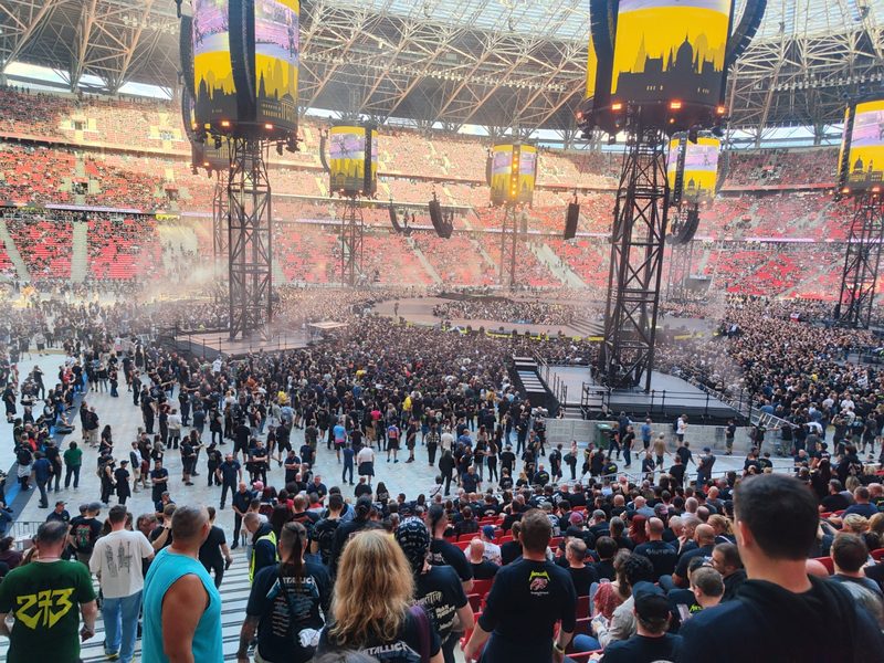 A stadium filling with people before a concert, with stage rigging and screens hanging above the floor.
