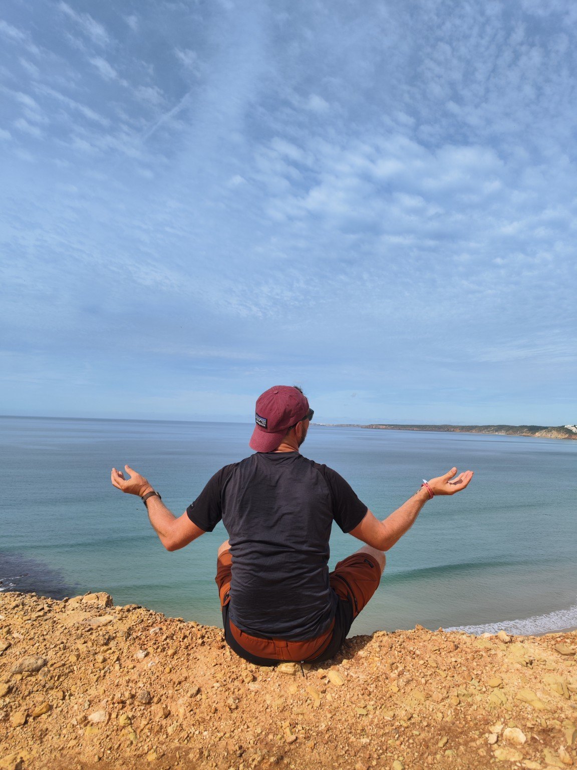 Sitting on a clifftop above the Atlantic on the Fishermen’s Trail in Portugal, seen from behind, arms open.